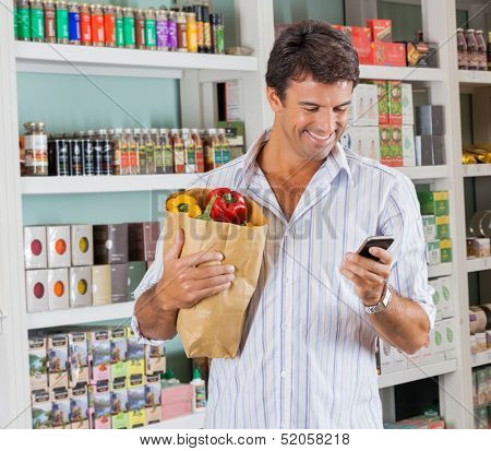 Happy male customer with grocery paper bag using mobile phone in supermarket