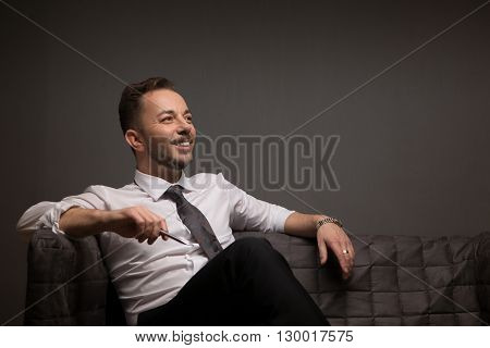 Handsome businessman sitting on sofa and looking away. Young man witout jacket resting and relaxing in studio.