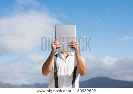 Young man covering face with digital tablet. Man hiding his face behind digital tablet while standing against clear sky. Portrait of a casual man holding digital tablet.