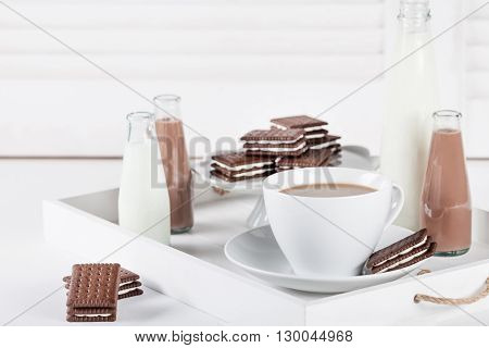 Cup of coffee and few bottles of milk and chocolate milkshakes on a white tray on white shutters background
