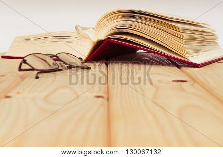 A pair of glassses on wooden table with an open book behind.