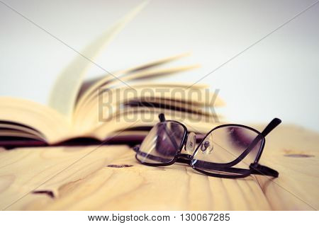 A pair of glasses on wooden table with an open book behind.