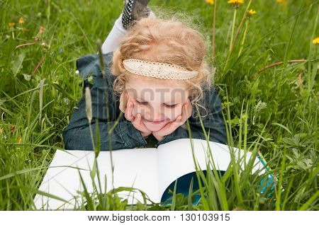 A little girl is lying on her stomach reading on the grass. She has a look of enjoyment on her face and she looks very relaxed.