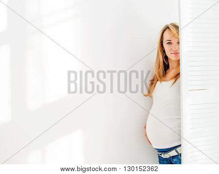Young and beautiful pregnant woman looks from behind a white screen on a background of a white wall in blue jeans and a white T-shirt