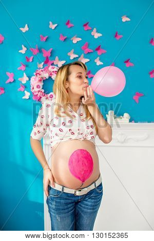 Happy pregnant woman blowing balloon against the backdrop of a blue wall with pink butterflies