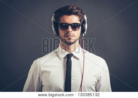Feel his beat. Portrait of handsome well-dressed young man in headphones looking at camera while standing against grey background