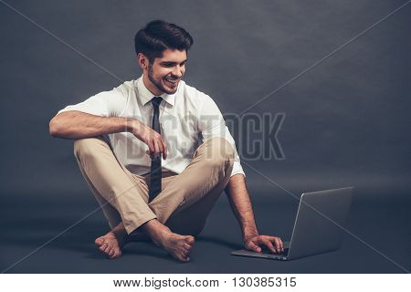 Staying online. Full length of confident young handsome man using laptop with smile while sitting on the floor against grey background