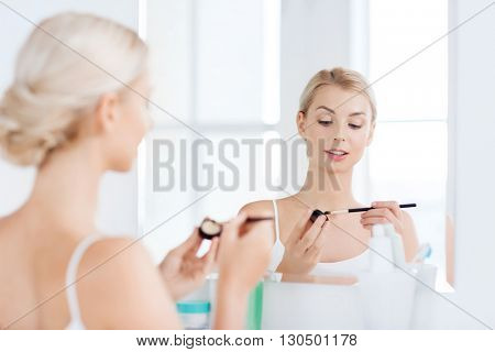 beauty, make up, cosmetics, morning and people concept - young woman applying eyeshade with makeup brush and looking to mirror at home bathroom