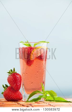 Glass of strawberry lemonade with pieces of strawberry, lemon and fresh mint. Closeup of mojito with beach on background. Fresh summer bright strawberry cocktail.