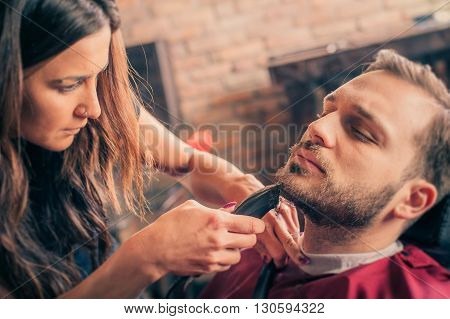 Female barber shaving a client's beard with trimmer in a barber shop