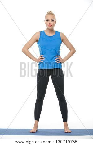 Cute blonde girl in the sportswear stands on a blue gymnastic mat on the white background in the studio. She wears black pants and blue sleeveless t-shirt. She is barefoot. She holds her hands on the waist. She looks into the camera with pouty lips. Verti