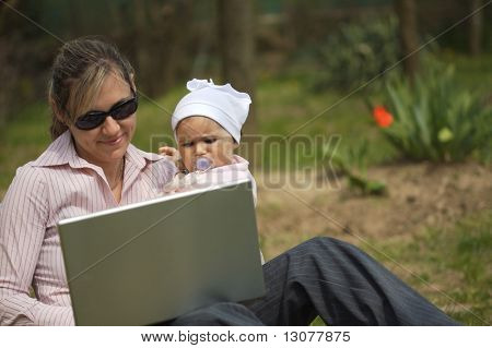 Mutter von einem 9 Monate alten Baby-Mädchen sitzt auf dem Boden im Garten und verwendet einen Laptopcomputer