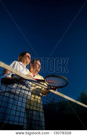 Aktiv senior par poserer på tennisbanen med tennisketsjer i hånden. Udendørs, sollys.