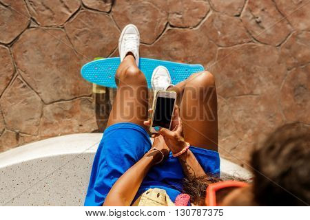 Beautiful Teenage Black Girl In Blue Skirt Sit With Her Smartphone.