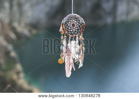 Handmade pink native american dream catcher on background of rocks and lake. Tribal elements feathers shells lace