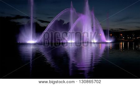 Amazing fantastic colorful fountain with bright illumination on the water pond or river with beautiful reflection at the evening or night. Beauty urban recreational concept.