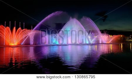 Amazing fantastic colorful fountain with bright illumination on the water pond or river with beautiful reflection at the evening or night. Beauty urban recreational concept.