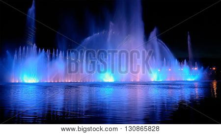 Amazing fantastic colorful fountain with bright illumination on the water pond or river with beautiful reflection at the evening or night. Beauty urban recreational concept.