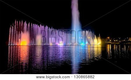 Amazing fantastic colorful fountain with bright illumination on the water pond or river with beautiful reflection at the evening or night. Beauty urban recreational concept.