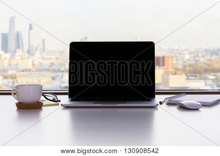Front view of blank laptop screen coffee cup and other items on white desktop with sunlit city in the background. Mock up