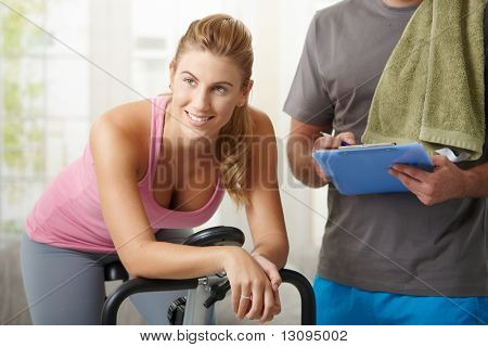 エアロバイクのパーソナル トレーナーと自宅でトレーニング、笑みを浮かべて若い女性。