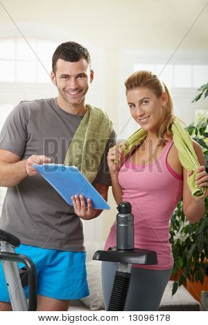 若い女性チェック トレーニング計画運動用の自転車の横にある自宅で立っている彼女のパーソナル トレーナーを。