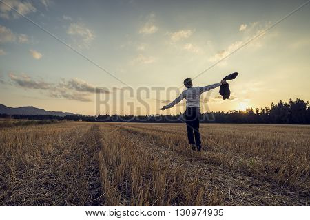 Man in elegant suit standing in the middle of cut field holding his jacket in on hand with his arms spread widely under an evening sky toned retro effect.