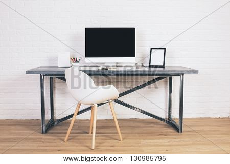 Front view of modern white chair and designer desk with blank computer screen and picture frames on white brick background. Mock up