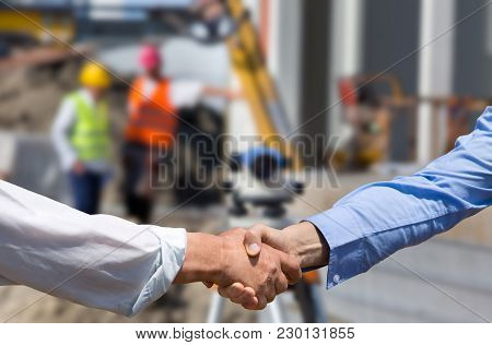 Two Business People Shaking Hands In Front Of Leveling Instrument On Construction Site With Workers 