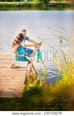 Family In Summer On A Picnic Near A Pond, Water. Family Holiday In Nature. A Little Daughter With He