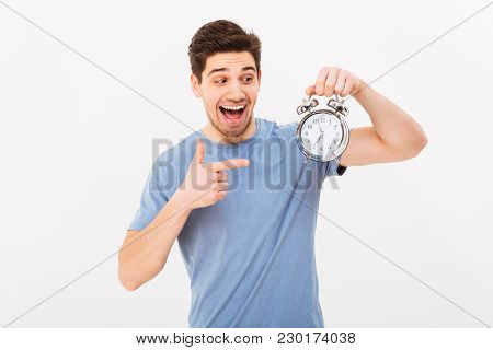 Young excited man in casual t-shirt and holding and pointing finger on alarm clock isolated over white wall