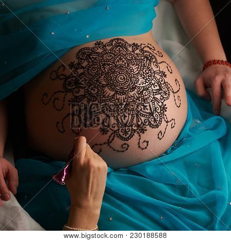 Mehendi artist paints an ornament of mandala with henna on an pregnancy woman’s belly