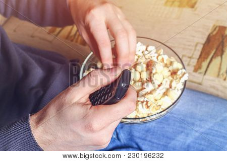 A Man With A Bowl Of Popcorn And A Remote Control In His Hand Looks At The Tv On The Sofa