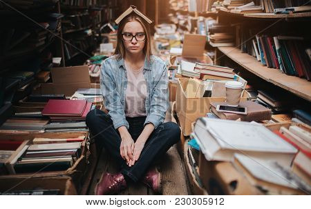 Attractive And Nice Girl Is Standing Among Big And Long Bookshelfs With Old Books. She Is Holding A 