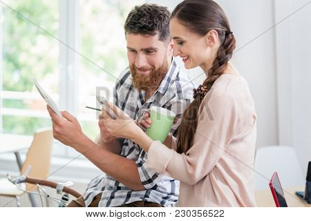 Two young co-workers smiling, while looking at a funny photo or watching a video on the mobile phone during break in a modern shared office space