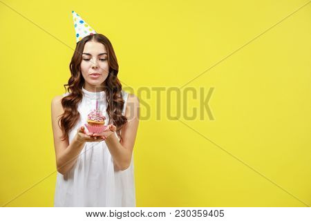 Beautiful young woman snuffing out candle on birthday cupcake against color background