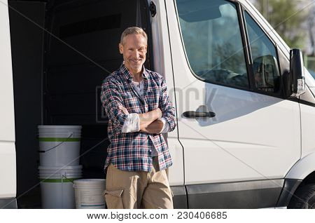 Portrait of painter standing next to his van with paint container. Decorator standing next to van and looking at camera. Happy smiling worker leaning on his white truck with folded arms.
