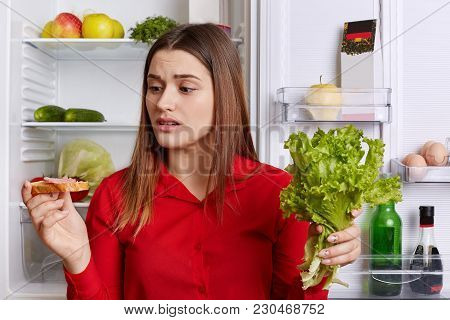 Horizontal Shot Of Brunette Female With Upset Expression Looks At Sandwhich, Keeps Lettuce, Wants To