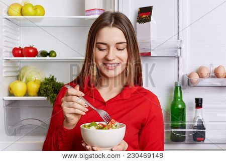 Horizontal Shot Of Cheerful Female Holds Bowl With Fresh Vegetable Salad, Stands Near Friedge Full O