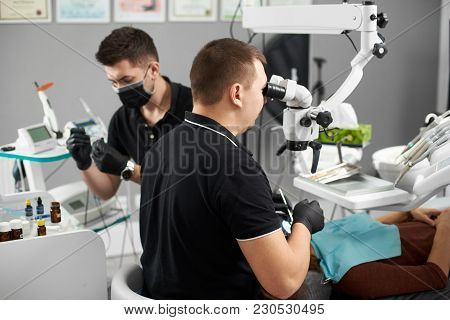Stomatologist With An Assistant In A Black Gloves Treats A Patient Using A Microscope. Modern Dentis