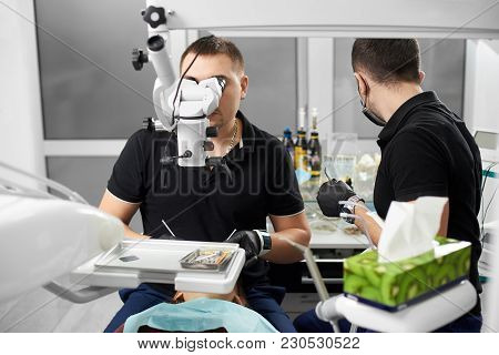 Man Dentist With An Assistant Treats A Patient Using A Microscope. Modern Dentistry With The Use Of