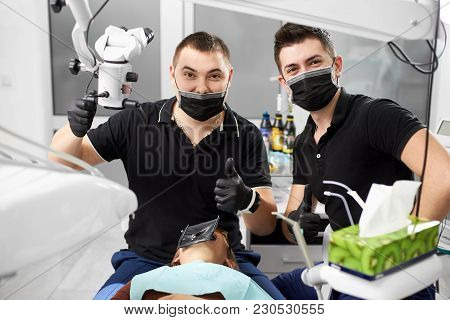 Young Dentist And His Assistant Wearing Black Masks, Gloves And Uniforms Are Looking Into The Camera