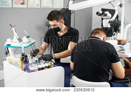 Young Assistant In A Black Mask And Gloves Helps The Dentist In The Process Of Treating The Patient.