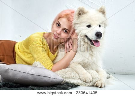 Attractive Young White-skinned Blonde Girl With Her Pet Samoyed Posing Lying On The White Floor And 