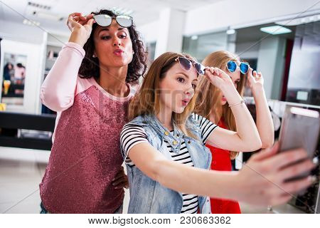 Three Young Stylish Girlfriends Raising Fashionable Sunglasses While Taking Selfie With Smartphone I