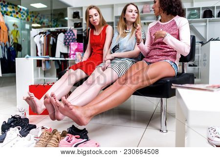 Young Smiling Girlfriends Sitting In A Clothing Store Looking At Their Bare Feet And Pile Of New Sho