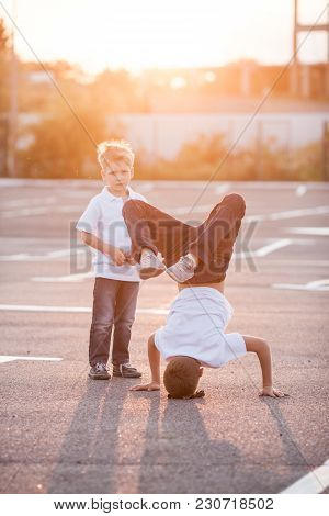 The Boy Is Showing A Headstand On The Street. Younger Brother Admires His Brother