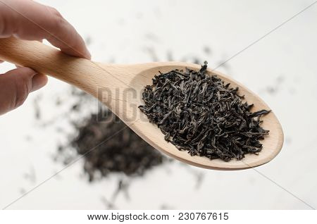 Hand Holds Dry Black Tea Leaves In Wooden Spoon, Scattered Tea Leaves Over White Background, Top Vie