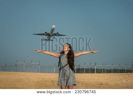 Jet Plane Takes Off On The Background Behind A Young Woman. Female Traveler Is Standing On The Beach