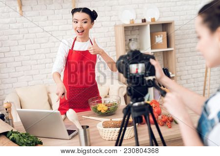 Two Culinary Bloggers In Red Aprons Read From Laptop With One Girl Behind Camera.